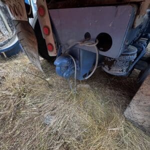 Close-up of a blue trailer hitch and electrical/air connector hanging from the underside of a trailer, with dry grass beneath and worn components nearby.