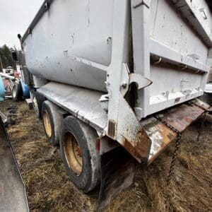 Rear side of a white, rusted dump trailer with multiple wheels, mudflaps, and a rusty tailgate in a grassy junkyard lot.