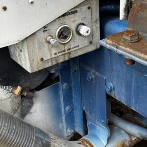 Close-up of a weathered industrial control panel with a gauge and two knobs mounted on a blue steel frame, showing rust and grime on surrounding components.