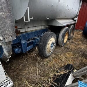 Side view of a grey tanker trailer with blue undercarriage and multiple wheels, parked on a grassy, muddy lot with a red building in the background.
