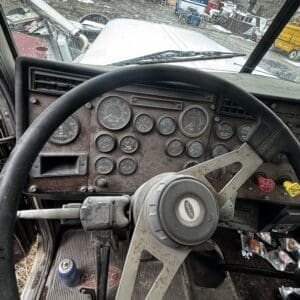 Interior of an old, dusty truck cab with a multi-gauge dashboard and large steering wheel; view out to a junkyard full of trucks and a red-roofed building.