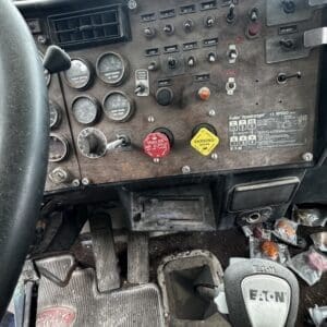 Dusty truck cab interior with analog gauges, switches on a metal dashboard, floor pedals, and an Eaton gear shifter.