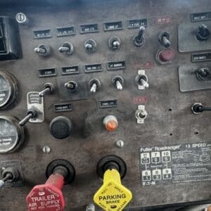 Dash panel with multiple switches and gauges, including a red trailer air supply knob and a yellow parking brake knob, in an old truck or tractor cab panel, worn metal surface.