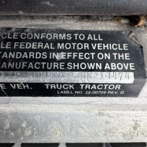 Close-up of a metal vehicle label: 'Truck Tractor', label No. 22-00709 Rev. G, certifying compliance with federal motor vehicle standards.