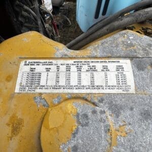 Close-up of a yellow Caterpillar engine housing with a worn chevron label listing engine model, displacement, and specs.