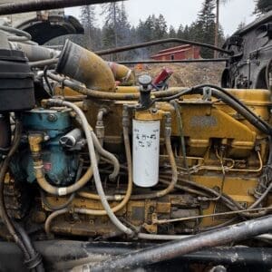 Diesel engine with yellow housing and exposed hoses, utility filter in foreground, outdoors with trees and a red shed in the background.