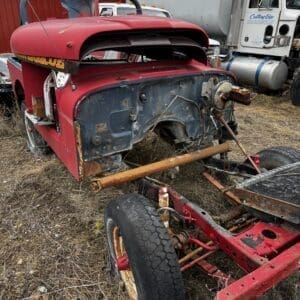 Rusty red vintage truck cab on a chassis in a junkyard, with an exposed engine bay and corroded metal parts in the grass.