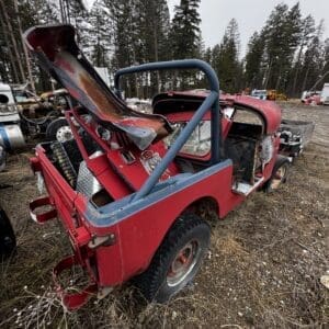 Red off-road jeep in a salvage yard, with a bent open hood, exposed engine bay, and a blue roll cage, surrounded by other junked vehicles and tall trees in the background.