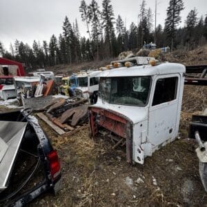 White truck cab with rusted front end in a junkyard, piled with rocks and debris, surrounded by abandoned vehicles and trees in the background.