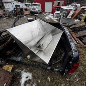 Metal industrial hopper resting in the bed of a pickup truck at a junkyard, with a red shed and old trucks in the background.