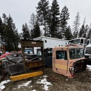Old truck cab and rusted machinery in a junkyard with other vintage vehicles and trailers behind.