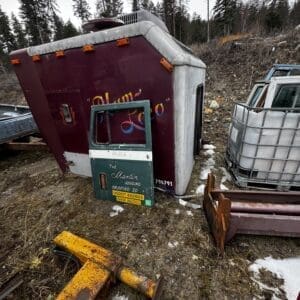Abandoned maroon and silver trailer tilted on its side in a junkyard with rusted equipment nearby in a forested lot.