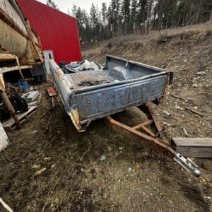 Rusty blue utility trailer with a hitch sits on a rocky, debris-strewn lot beside a red metal building, near a forested slope.