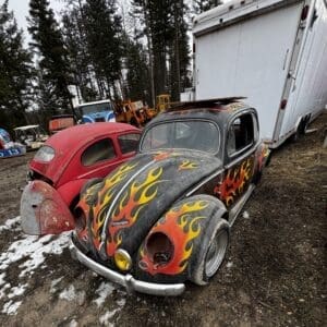 Black vintage VW Beetle with yellow-orange flame decals parked beside a red car in a junkyard with a large white trailer behind them.