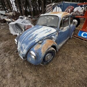 Blue vintage VW Beetle with heavy rust and dirt sits in a junkyard among scattered salvage and tarps.