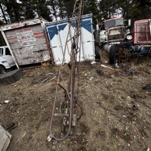 Rusty metal derrick/drilling rig standing upright in a junkyard with abandoned trucks and trailers in the background.