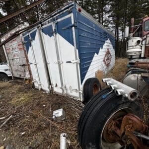 Weathered blue-and-white cargo trailer tilted in a junkyard, with rusted wheels and tall grass around it.