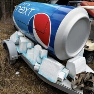 Blue Pepsi Next cylindrical tank on a trailer, surrounded by white foam packing blocks in a wooded area.