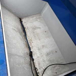 Inner metal trough or cabinet with a blue pipe secured to the floor and a black electrical cable, dirty concrete base and blue walls nearby.
