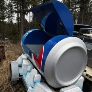 Blue cement mixer on a trailer with its drum open, surrounded by white foam blocks in a rural, wooded area with a pickup truck nearby.