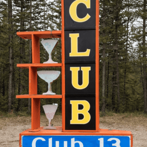 Large orange frame sign with black panels and yellow letters spelling CLUB, with three decorative cocktail glasses on orange shelves, set in a forested area; base reads Club 13.