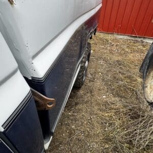 White pickup truck with a large dent along the passenger side and a rusted metal bracket protruding from the gap, on dirt near a red wall.