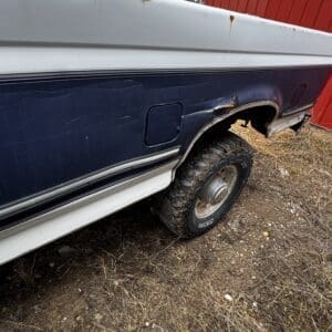Rear side of a two-tone blue and white pickup with rust spots and a damaged wheel well, parked on dirt near a red shed.