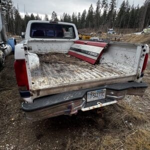 White Ford pickup with the bed down, dirty and filled with debris, parked on a muddy, forested construction site.