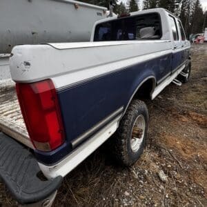 Rear three-quarter view of a blue-and-white pickup truck with a dented tailgate, parked on a gravel lot beside a trailer in a wooded area.