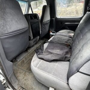 Dusty pickup truck backseat with worn gray upholstery and a rumpled blanket/piece of clothing on the seat, view of dusty floor mats and a plastic bottle on the floor behind the driver’s seat