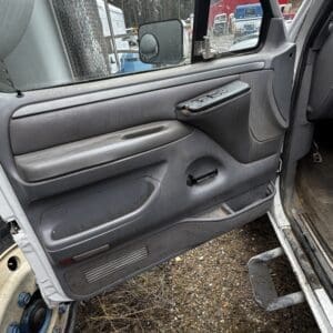 Open truck door interior: gray door panel with armrest and power window controls, yard with trucks visible outside.
