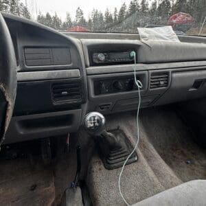Dusty vehicle interior with a 5-speed manual shifter, center console, and plugged-in stereo; green charging cable drapes across the dashboard.