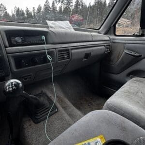 Inside a dusty car cabin with a manual gear shift, dashboard controls, and a hanging cord; forested trees visible through the windshield.