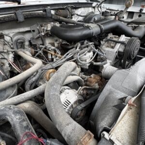 Open car hood exposing a dusty engine bay with hoses, belts, and an alternator visible among rust and grime, viewed from above.