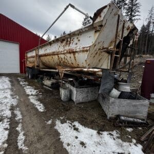 A large, rusted white dump trailer parked on a muddy yard beside a red metal building, with snow patches and scattered parts underneath and around it.