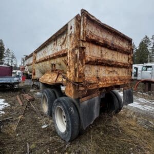 Rusty, weathered dump truck trailer in a junkyard with other old vehicles nearby.