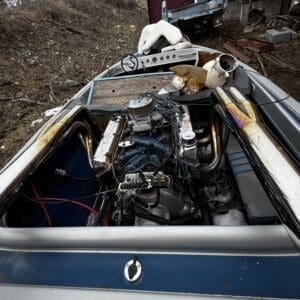 Old speedboat with its motor exposed, in a junkyard among rusted vehicles and scrap on a dirt lot.