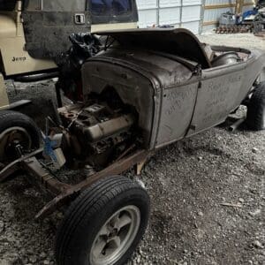 Rusty vintage open-top roadster with exposed engine on a gravel lot, next to a dusty Jeep and a large metal storage building in the background.