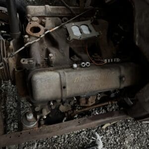 Rusty vintage car engine mounted in a bare chassis, with exposed manifolds and fuel lines.