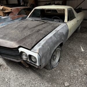 Vintage mid-century car in poor condition with a rusted body and missing hood, parked inside a metal shed.