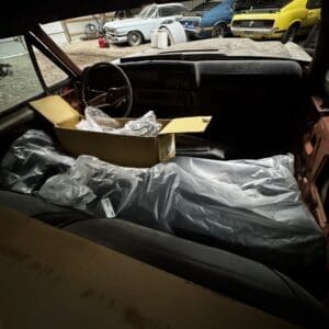Interior of an old car with a cardboard box and plastic bags on the passenger seat, garage clutter visible outside (classic cars in background).