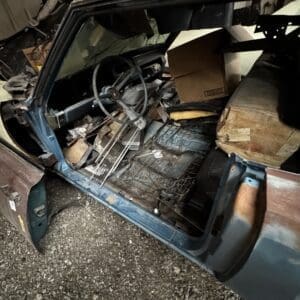 Open driver’s door of a blue, rusted truck interior cluttered with boxes, debris, and a metal rack on the floor.