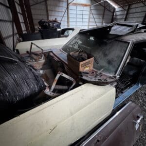 Old, dusty vintage car inside a cluttered metal shed, with a cardboard box and debris on the hood.