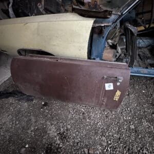 Detached brown car door resting on gravel in a cluttered garage, with an old two-tone blue and beige car body in the background and various parts around it.