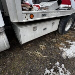 Utility truck bed with scattered metal scraps, hoses, and a silver storage box on the ground under a dump truck chassis.