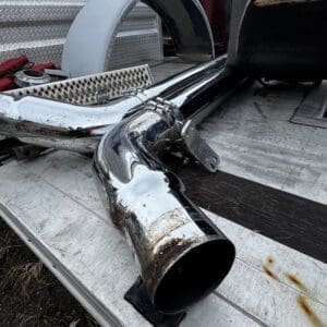 Chrome truck exhaust pipe and other metal components resting on a weathered flatbed with diamond-plate surface; visible rust and wear.