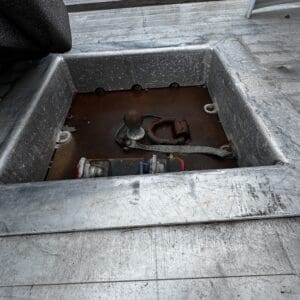 Open hatch on a metal boat deck showing a rusted ball hitch, winch, and rope/strap inside a recessed compartment.