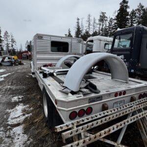 Flatbed trailer with large curved wheel fenders on a snow-dappled, muddy lot; rusty metal beams stacked nearby and trucks in the background in a wooded area.