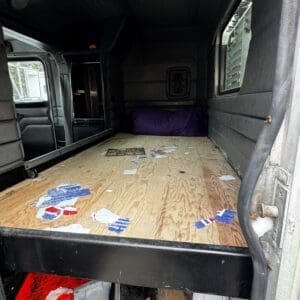 Cargo van interior with a plywood floor, cluttered with scraps and a purple cushion at the far end.