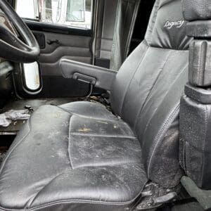 Interior of a dusty, well-worn pickup truck cab with a black leather driver’s seat and dirty floor mats; steering wheel and door panel visible. Outside view shows another truck cab through the window.